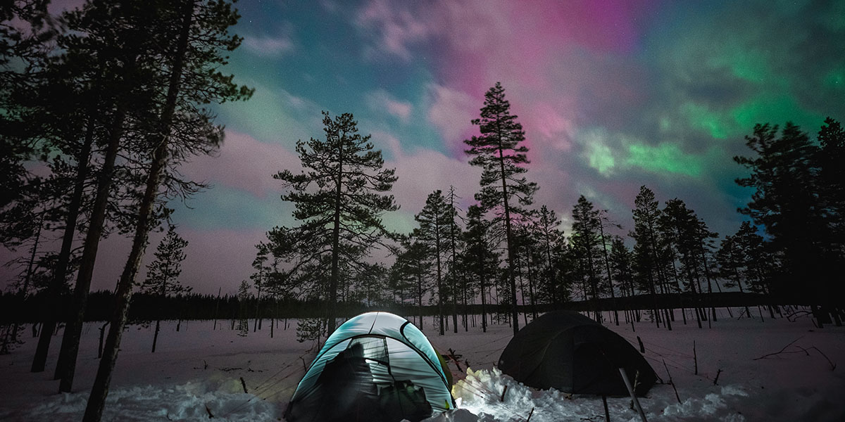 Zwei Zelte im Schnee, unter einem leicht mit Wolken bedeckten Himmel, welcher von Nordlichten erleuchtet wird.