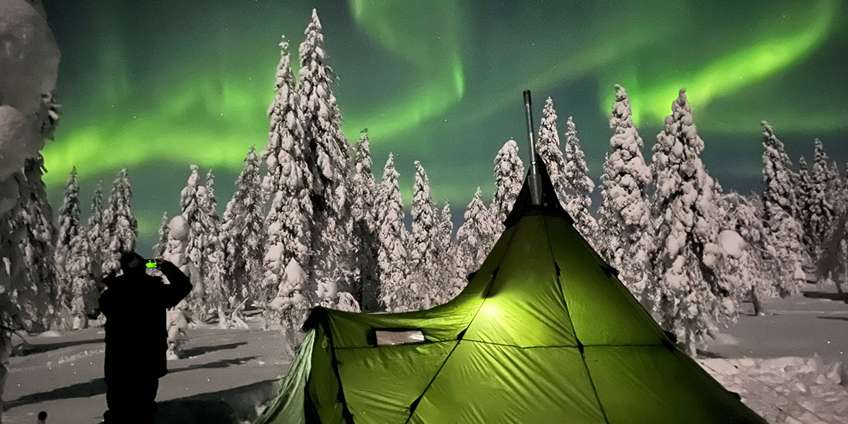 Eine Nordlichter fotografierende Person die neben einem grünen Zelt im Wald steht und im Hintergrund Schnee bedeckte Bäume mit einem dunkelblauen Himmel und grünen, tanzenden Nordlichter darüber.
